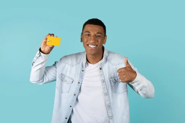 快乐的年轻非洲裔美国人穿着休闲装 带着信用卡和大拇指 推荐网上购物 被蓝色背景隔离 工作室 大甩卖 折现等方面的建议 — 图库照片