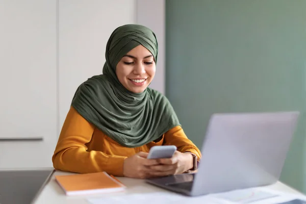 在希吉布用智能手机微笑年轻的穆斯林妇女在办公室里 快乐的伊斯兰女性坐在电脑前 在手机上留言 享受在线交流 自由自在的空间 — 图库照片