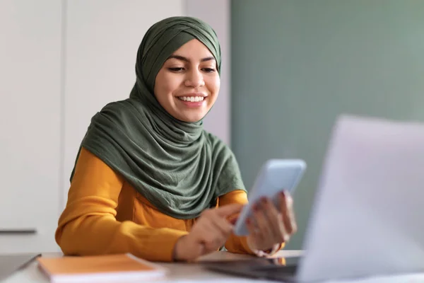 在Hijab用智能手机在线购物的穆斯林妇女坐在办公桌前 快乐的年轻伊斯兰女士在手机上浏览应用程序 喜欢在网上购物 闭门造车 — 图库照片