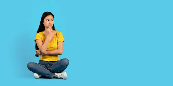 黄色のTシャツの深刻な百周年記念韓国の女性の学生の床に座って 空の空間を見て 考えて 青の背景 スタジオに隔離されます 勉強や仕事 人々の感情のための広告と提供 — ストック写真