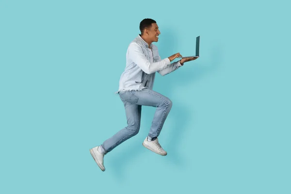 Happy busy young african american man student or manager typing on laptop, chatting, jumped, frozen in air isolated on blue background, studio. Technology for work, study, deadline and new normal