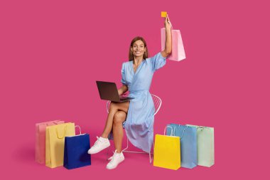 Gülümseyen, kafkasyalı, bol çantalı, dizüstü bilgisayarı olan, pembe arka planda, stüdyoda izole bir şekilde oturan sarışın bir kadın. Reklam, teklif, büyük indirim, online alışveriş, nakit para.