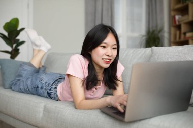 Evden çalışan Asyalı genç bayan serbest yazar, kanepeye uzanıp modern dizüstü bilgisayar kullanıyor, müşterilerle internet üzerinden çalışıyor veya iletişim kuruyor, blog yazıyor, kopyalama alanı kopyalıyor.