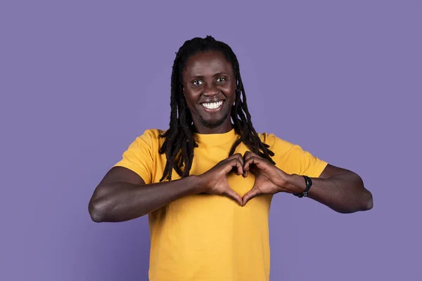 胸と笑顔の上にハート型のシンボルを示す黄色で遊び心のあるアフリカ系アメリカ人の若い長髪の男 紫のスタジオの背景に愛と忠誠心を示し 感情を共有し スペースをコピー — ストック写真