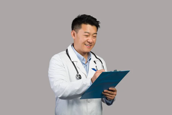 快乐的日本成年男性治疗师 身穿白衣 带听诊器 在平板上做笔记 在灰色背景下隔离 工作室 诊所的医生工作 体格检查 保健和治疗 — 图库照片
