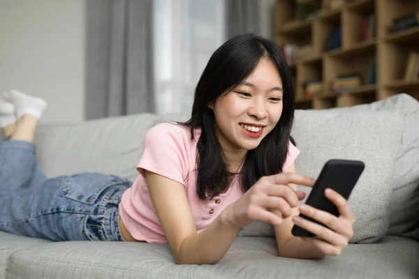 アジアの女性は ソファに横たわって 自宅でリラックスし 携帯電話でニュースを見て 一人で暇な時間をお楽しみください 若い韓国の女性のスマートフォンでテキストメッセージ リビングルームのインテリアでインターネットサーフィン — ストック写真