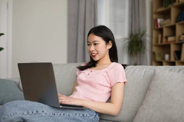 幸せな韓国人女性のフリーランサーはソファに座って ノートパソコンのキーボードと笑顔を入力し 電子メールやサーフィンをインターネット 家庭内 無料のコピースペースで送信します — ストック写真