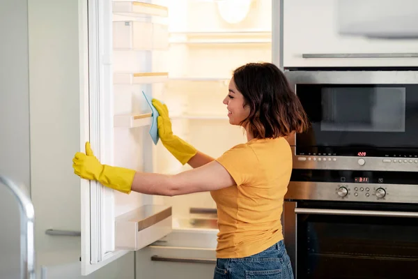 若いアラブ女性は 台所で空の冷蔵庫をきれいにするためにラグを使用して ゴム手袋を身に着けている陽気な中東の女性は家にタイディング 笑顔の女性は家事の雑用を楽しむ フリースペース — ストック写真