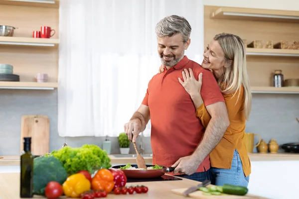 家庭的な概念で料理 幸せな感謝美しいブロンド中年女性抱擁彼女の夫後ろから 愛する成熟した白髪の男準備健康的な食事 居心地の良いキッチンインテリア コピースペース — ストック写真