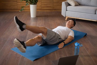 Olgun Koreli bir adam oturma odasının iç kısmında paspas ve karın kasları için egzersiz yapıyor ve dizüstü bilgisayarda antrenman izliyor. Çevrimiçi antrenör, kilo kaybı ve vücut bakımı