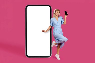 Beyaz elbiseli, kablosuz kulaklıklı mutlu, beyaz kadın akıllı telefon, pembe arka planda, stüdyoda izole edilmiş, boş ekranlı, dev bir telefonun yanında dans ediyor. Reklam ve teklif, bayan müziğin tadını çıkarın, uygulama