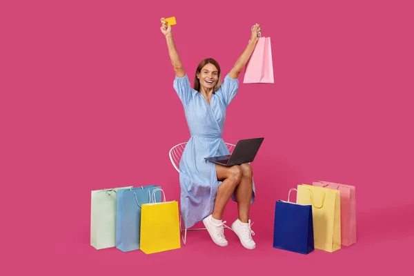 快乐而震惊的成年高加索金发女性 穿着多包扎的衣服 手持笔记本电脑 手持信用卡 享受成功的网上购物 与粉色背景隔离 — 图库照片
