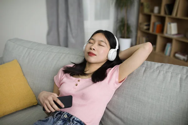 ソファの上に寝そべって家で目を閉じて音楽を聴くワイヤレスヘッドフォンの若いリラックスしたアジアの女性 穏やかな韓国の女性は好きなメロディーを楽しんで — ストック写真