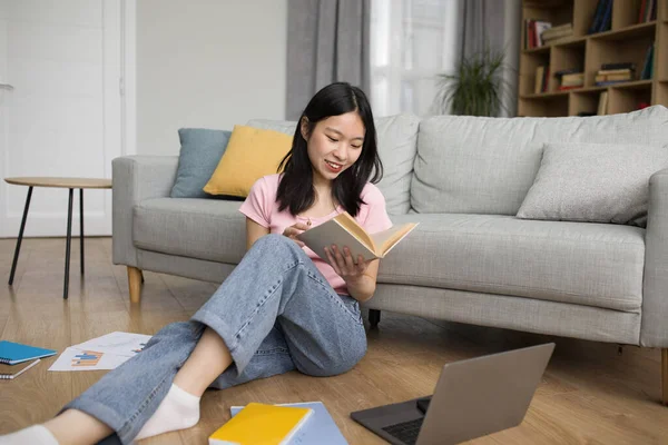 Happy young japanese lady studying online from home, reading book and using laptop pc, sitting on floor in living room, free space. E-learning, web-based education