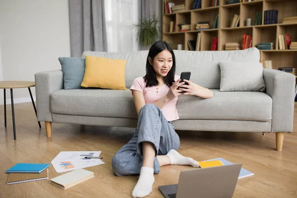 幸せな韓国人女性は自宅でノートパソコンやスマートフォンでリラックスし リビングや携帯電話でのメッセージで床に座り オンライン通信を楽しんでいます — ストック写真