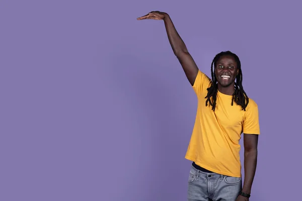 彼の手と笑顔で見えない何かの高さを示す黄色のTシャツで陽気なハンサムな長髪の若いアフリカ系アメリカ人男性 紫色のスタジオの背景にポーズ コピースペース — ストック写真