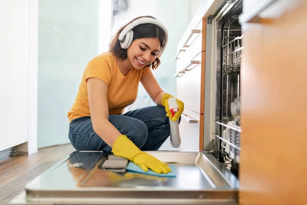 Happy Arab Female Cleaning Washing Machine In Kitchen With Rag And Sprayer, Cheerful Young Middle Eastern Woman Wearing Wireless Headphones Tidying Home And Listening Favorite Music, Free Space