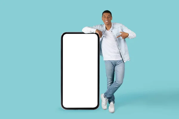 用随意的手指用空白屏幕对着巨大的手机微笑着的年轻的非洲裔美国人 推荐用蓝色背景隔离的新小玩意 应用演示 广告和提供的建议 — 图库照片