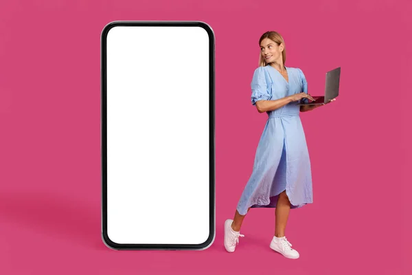 Smiling pretty adult caucasian blonde woman in dress with laptop looks at huge smartphone with blank screen, isolated on pink background, studio. Ad and offer, app for blog, study, work and business