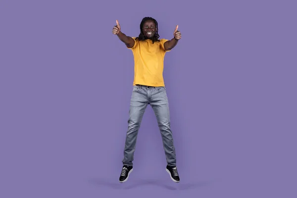 紫色のスタジオの背景に空気中でジャンプしながら親指を示すカジュアルな服装で幸せなハンサムな長髪の若い黒人男性 何かまたは誰かのような スペースをコピー 人間のジェスチャーの概念 — ストック写真