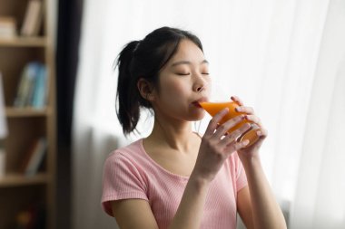 Susamış genç Çinli kadın camdan taze portakal suyu içiyor, evdeki pencerenin yanında duruyor, boşluğu kopyalıyor. Sağlık hizmeti, evde düzgün beslenme, kahvaltı ve günaydın.