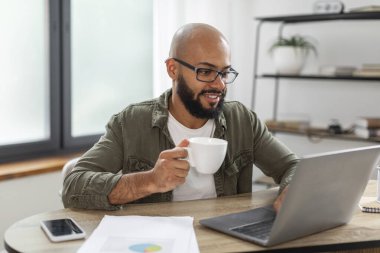 Laptop kullanan, elinde fincan tutan ve evde çalışırken sabah kahvesini içen pozitif Latin sakallı bir adam. Masada oturan ve bilgisayar kullanan serbest çalışan bir erkek.