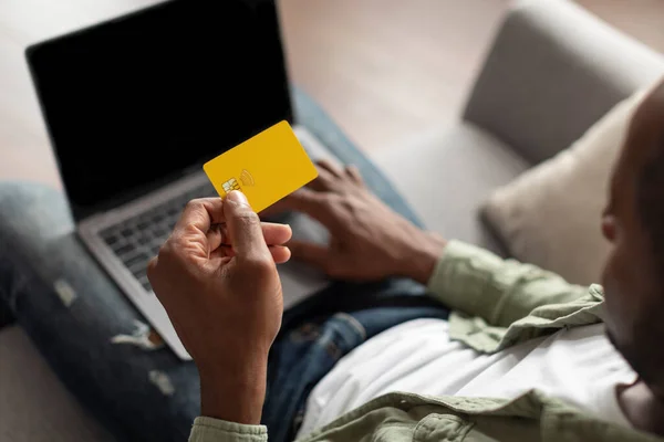 Adult black male typing on computer keyboard with empty screen, using credit card for online shopping and banking in living room interior. Financial management remote, purchase and order of goods