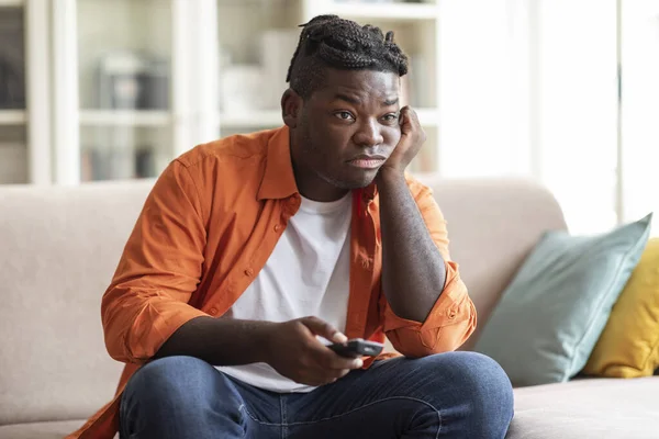 Boring overweight african american millennial guy with dreadlocks hairstyle sitting on couch at home, switching tv for fascinating film or show, free space. Domestic entertainment concept