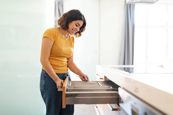 Young arab woman tidying up cutlery in drawer while cleaning in kitchen at home, happy middle eastern female assembling forks, enjoying making domestic chores, side view with copy space