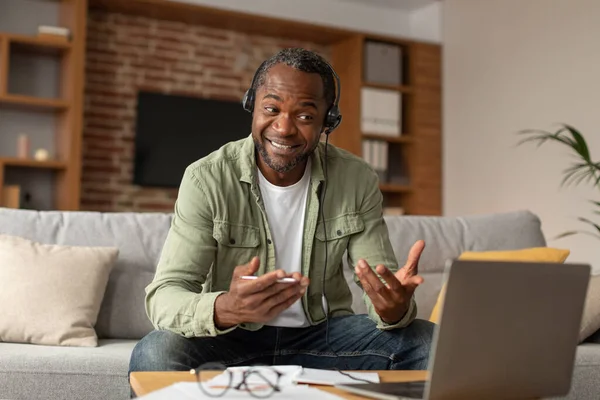 ヘッドフォンで幸せな中年の黒人男性は コンピュータ上のビデオ通話を持っており リビングルームで顧客をサポートしています 社会的距離 現代的な技術で自宅でリモート 仕事やビジネスを満たしています — ストック写真