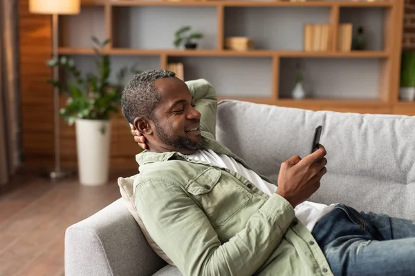 幸せな成熟した黒人男性は スマートフォンを使用して 時計のビデオは リラックスしたサーフィン ニュースを読む 自由な時間を楽しむ リビングルームのインテリアでソファの上に横たわる コピースペースを持っています 新しいアプリケーション 自宅でのチャットやソーシャルネットワーク — ストック写真