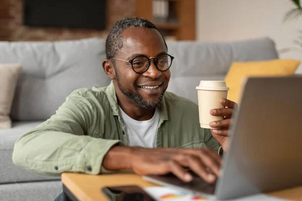 自宅で仕事やビジネス 幸せな大人の黒の男性の眼鏡をコンピュータ上で入力し リビングルームのインテリアでコーヒーをテイクアウトのカップをお楽しみください 閉じます 社会的距離と新しい通常のための近代的な技術 — ストック写真