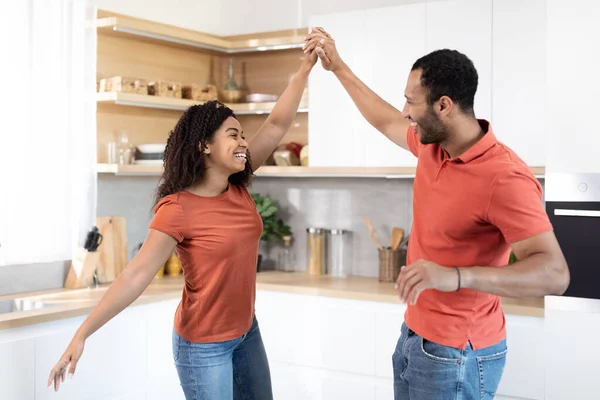 Cheerful millennial black couple in red t-shirts dancing, have fun together at music in kitchen interior, free space. Relationship, romance, love and date, family enjoy weekend and spare time at home