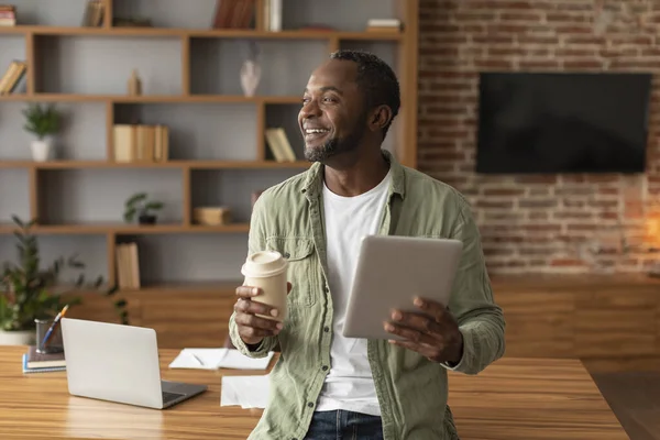 タブレットとコーヒーを飲みながら大人の黒人男性を笑いながら 自宅のオフィスのインテリアで無料のスペースで見てください 仕事とビジネス 近代的な技術とデータ分析 新しいアプリで休憩と休息 — ストック写真