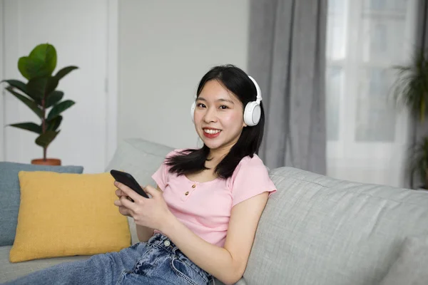 ソファに座って 自宅でスマートフォンを使って音楽を聴いているヘッドフォンの幸せな日本人女性は カメラで笑っています 若いですアジアの女性楽しみます彼女のお気に入りの歌やオーディオブック — ストック写真