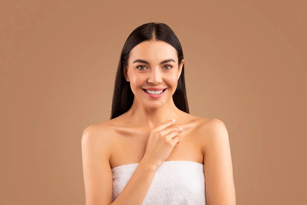 Body care concept. Half-naked beautiful long-haired brunette young woman wrapped in bath towel touching her skin, smiling at camera isolated over beige background, studio shot, copy space for advert