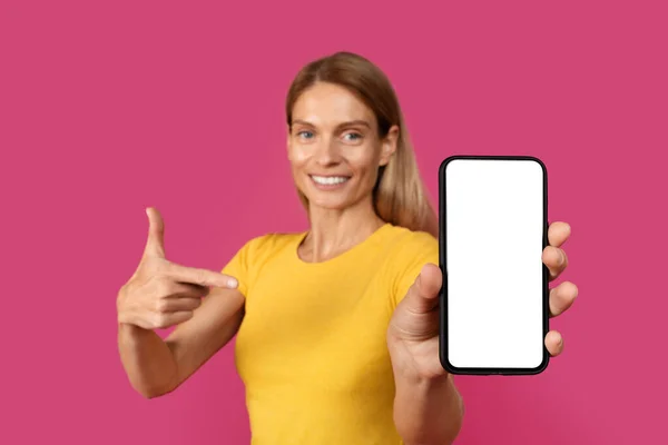 幸せな大人の白人女性のブロンドピンクの背景 スタジオに隔離された新しいアプリやデバイスをお勧めします空の画面でスマートフォンでカジュアルなポイント指で ウェブサイトの広告と提供 ブロガーのアドバイス — ストック写真