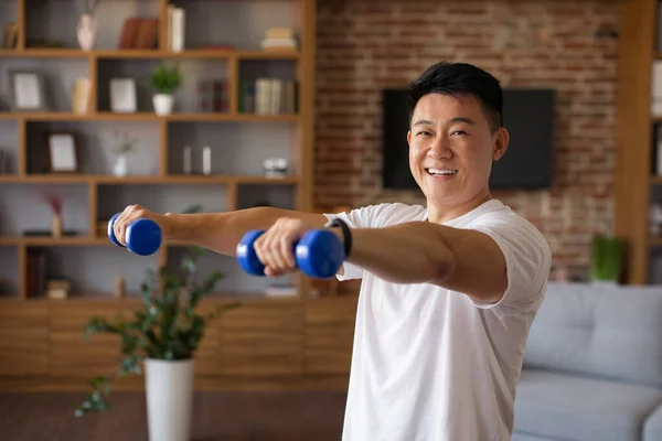 Home workout concept. Happy mature asian man doing domestic training with dumbbells, strengthening his biceps. Fit middle aged male taking care of his body, leading active lifestyle