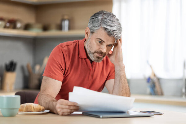 Crisis 2022. Unhappy middle aged male entrepreneur reading correspondence while having breakfast at home, sad man sitting at kitchen desk with laptop and smartphone on, holding documents, copy space