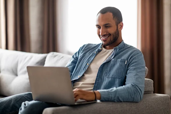 Online Correspondence. Smiling Black Man Sitting On Couch And Typing On Laptop, Happy Young African American Male Using Computer For Remote Work Or Communication, Resting On Sofa In Living Room