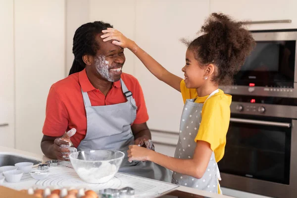 Cheerful African American Dad And Daughter Having Fun While Baking In Kitchen, Cute Preteen Black Girl Playfully Touching Fathers Face With Flour And Laughing, Enjoying Spending Time Together