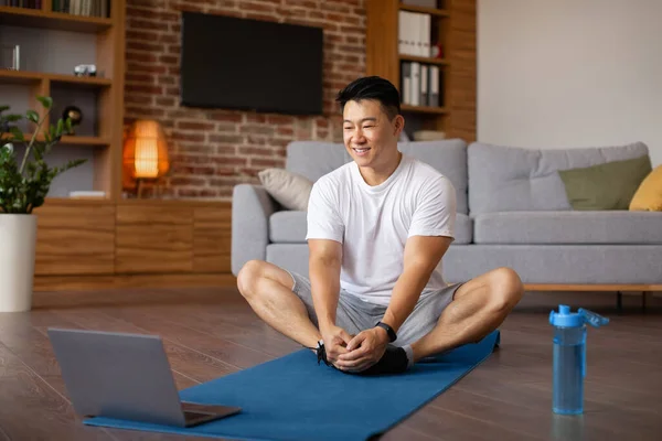 Online yoga practice. Mature asian man doing butterfly asana on sports mats, following video tutorial on laptop pc, copy space. Positive korean male exercising at home