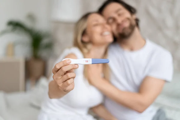 Happy caucasian spouses showing positive pregnancy test and embracing at home, selective focus. Loving husband and wife feeling excited to become future parents. Family planning concept