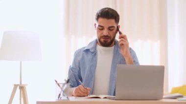 Uzaktan çalışma konsepti. Orta Doğulu genç bir adam müşterisiyle telefonda konuşuyor ve not alıyor, bilgisayarla evden çalışıyor, görüntüyü takip ediyor, yavaş çekim, serbest alan