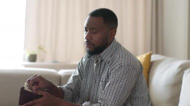 Genç Afro-Amerikalı adam cüzdanında para sayıyor, ev harcamaları ve iç bütçe planlıyor, mali istikrar konusunda kararsız hissediyor, izleme çekimi, yavaş çekim, serbest alan
