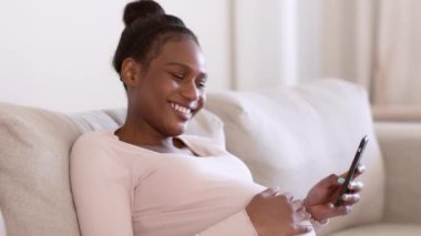Genç mutlu hamile Afrikalı Amerikalı kadın sosyal medyada sörf yapıyor, akıllı telefondan haberleri okuyor ve karnını okşuyor, evdeki kanepede dinleniyor, çekim takip ediyor, yavaş çekim, serbest alan.