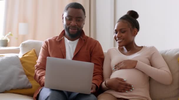 产前网上购物 年轻快乐的怀孕的非洲裔美国妇女和她的丈夫在笔记本电脑上冲浪 分娩前选择孩子 跟踪镜头 慢动作 自由自在的空间 — 图库视频影像