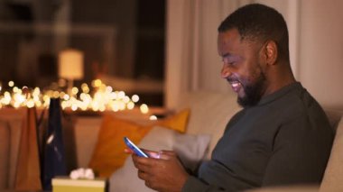 Çevrimiçi iletişim kavramı. Genç, kaygısız Afrikalı Amerikalı adam sosyal medyada arkadaşlarıyla sohbet ediyor, akıllı telefondan mesaj atıyor, akşamları evde dinleniyor, çekimlerin izini sürüyor, yavaş çekim, boş alan.