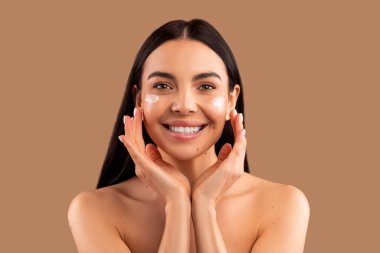 Yaşlanma önleyici göz bakım kozmetik konsepti. Mutlu, çekici, esmer, uzun saçlı, yarı çıplak, cilt kremi sürmüş, bej rengi stüdyo arka planı, fotokopi alanı.