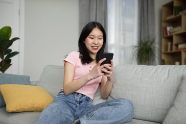Güzel alet ve başvuru formu. Cep telefonu tutan mutlu Asyalı kadın, mesaj yazıyor, oturma odasında kanepede oturuyor. Genç Koreli kadın internette geziniyor, internette sörf yapıyor, uygulama kullanıyor